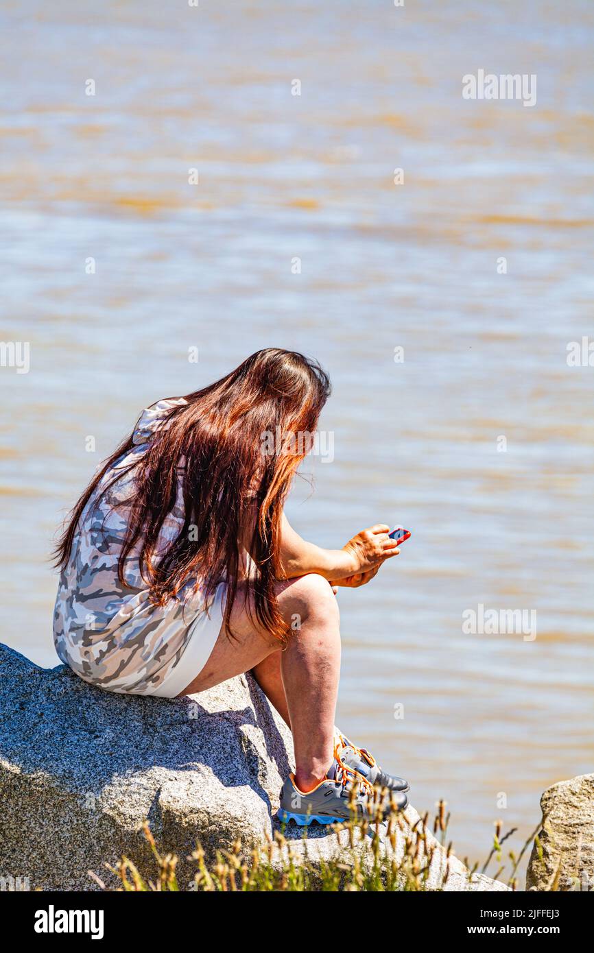 Young woman looking at her phone sitting on the edge of the Fraser River on Canada Day 2022 Stock Photo