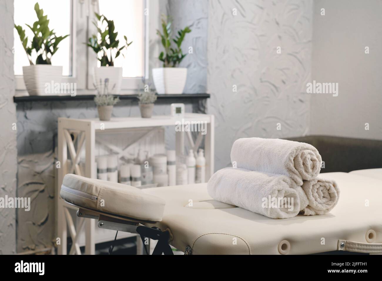 Rolled white towels on massage table in empty salon. Interior of aesthetic clinic, nobody. Modern cabinet of physiotherapist. Empty room with plants Stock Photo