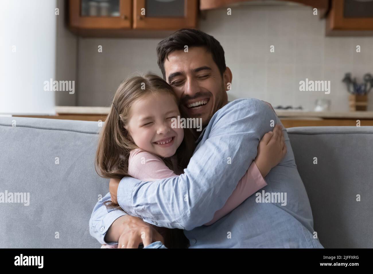 Little daughter cuddles tightly her stepdad seated on sofa Stock Photo