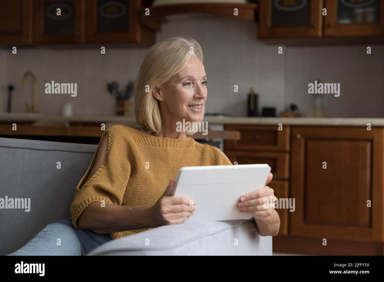 Smiling attractive older woman sit on sofa use digital tablet Stock Photo