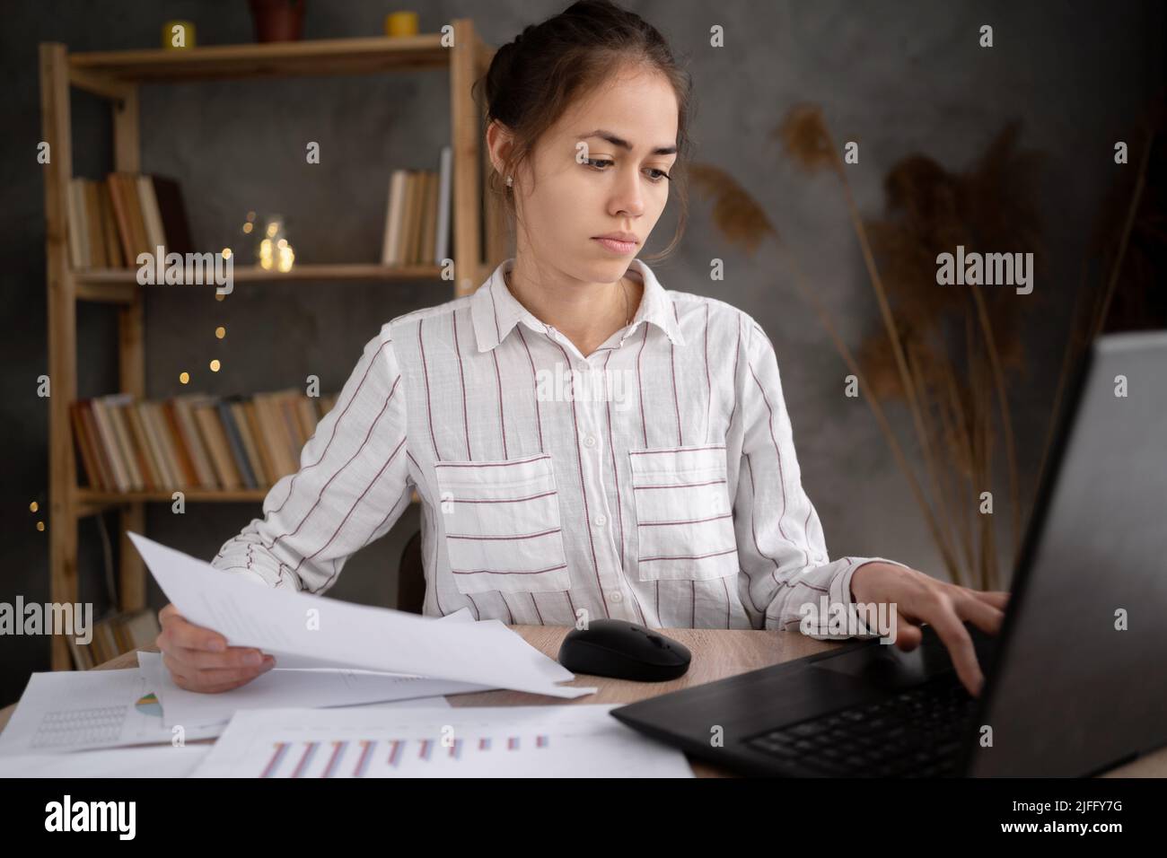 Portrait of serios hispanic young woman with laptop computer and papers working at home office. remote job, technology and people Stock Photo