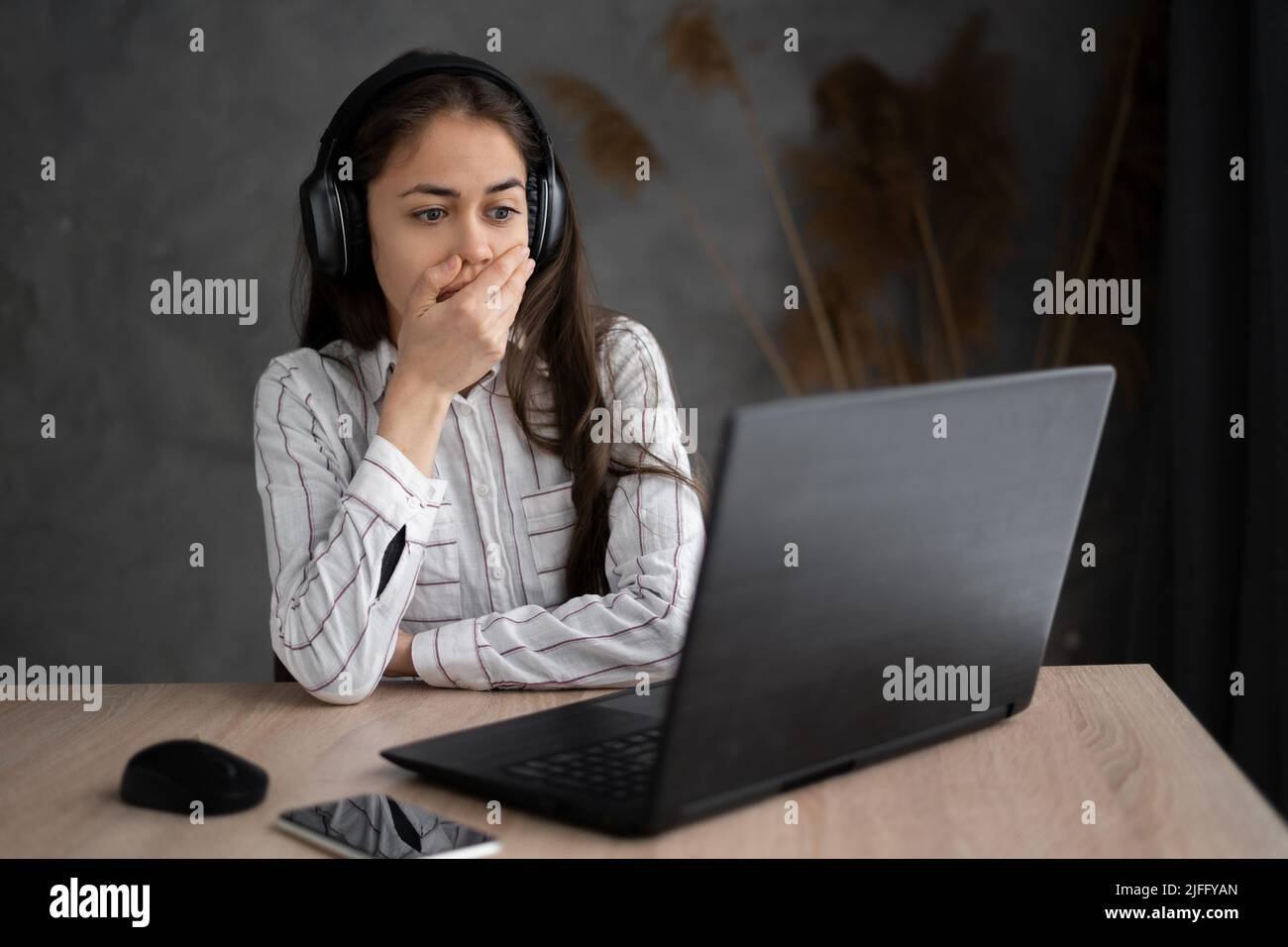 Portrait of shocked young woman watching horror film at home using headphone and laptop. She is covering her mouth by hand with surprise while staring Stock Photo