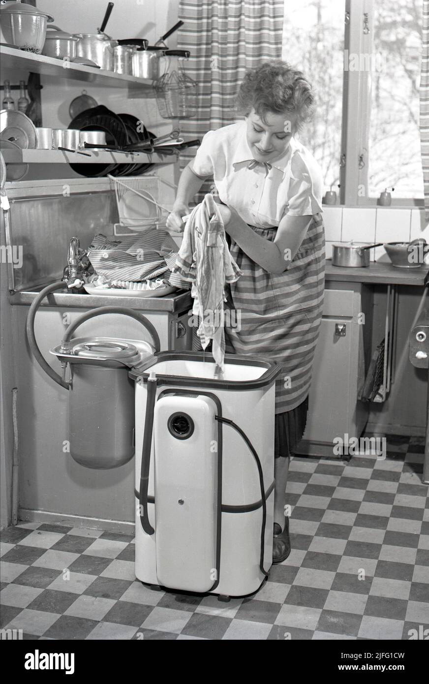 Doing the laundry in the 1950s. A lady is using a washing machine in the kitchen. A typical 1950s kitchen with a sink and pots and pans on the shelves above.  Sweden 1953 ref 2317 Stock Photo