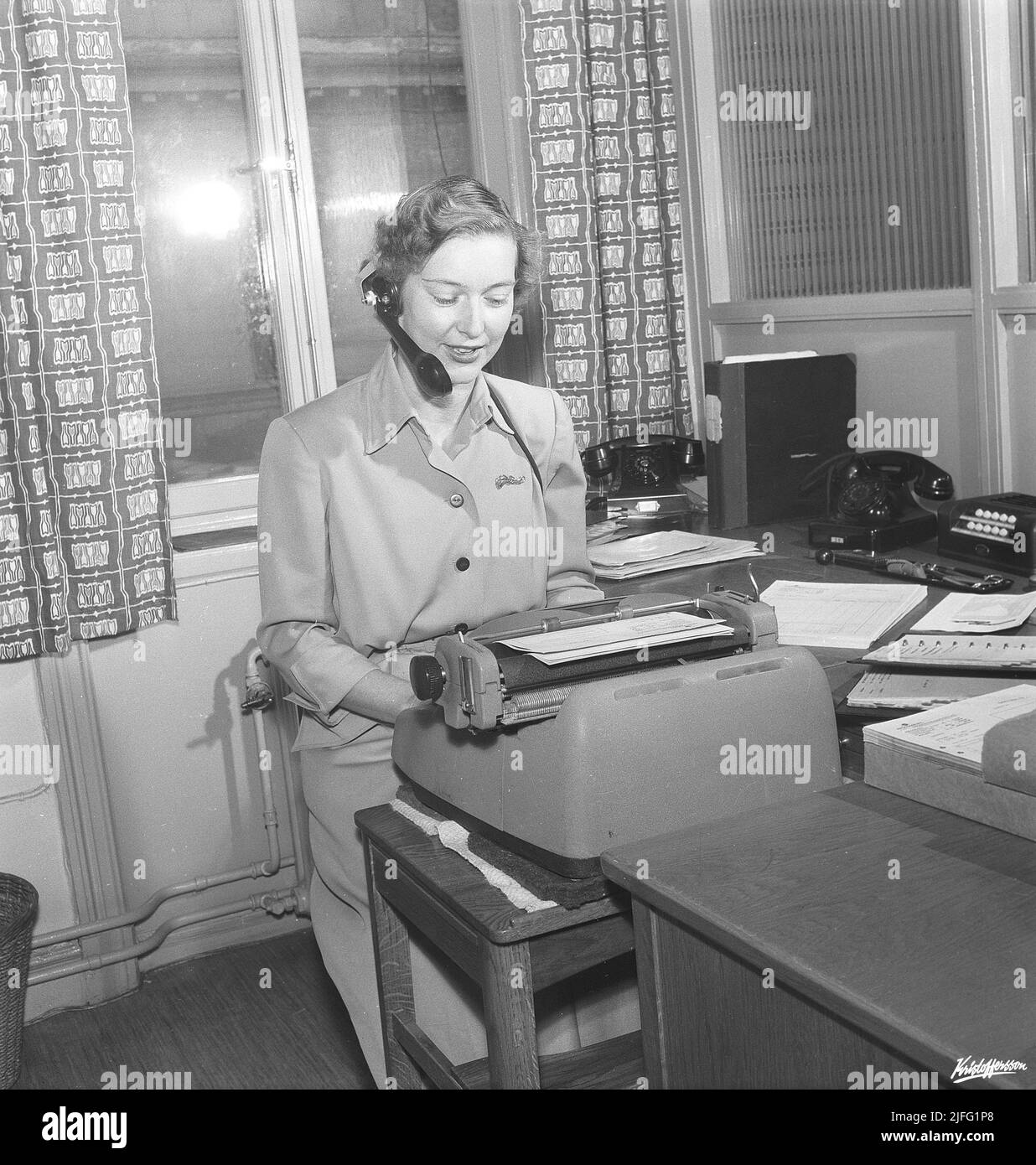 Office girl in the 1950s. A woman is sitting in front of her typewriter in an office, typing something in copies. She has a headset on, possibly listening to a recording of a letter or a message to type. Sweden 1955. Kristoffersson BM71-12 Stock Photo