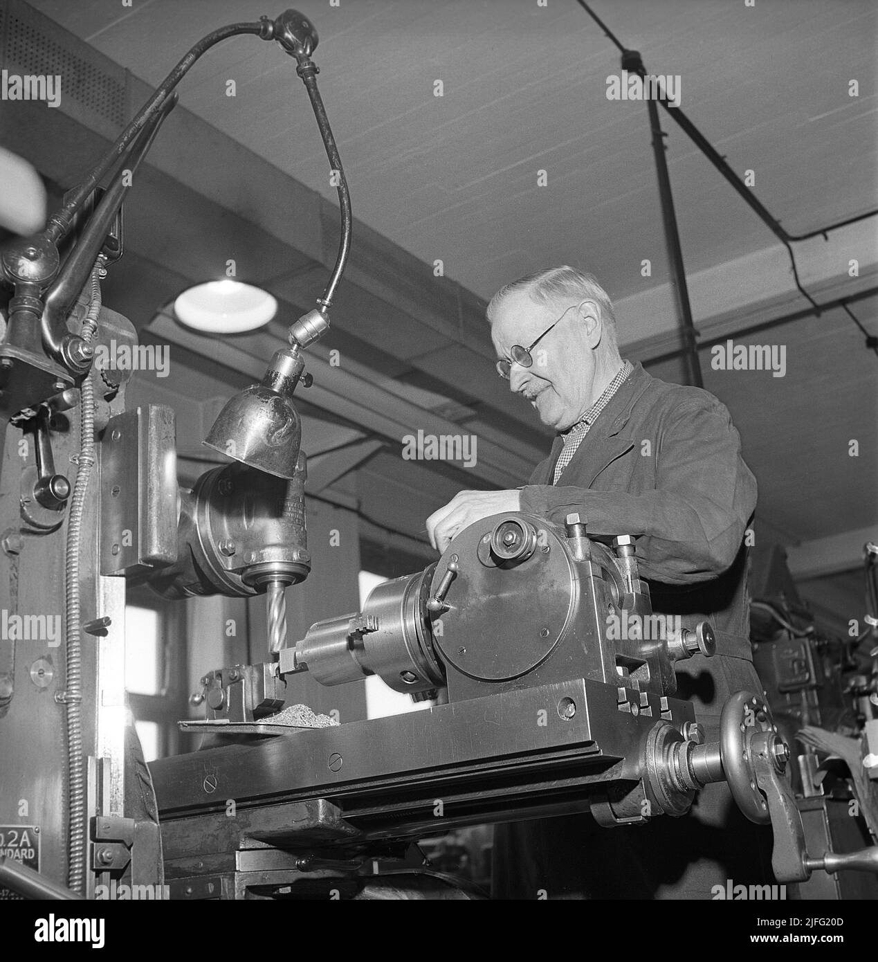 Mechanical workshop in the 1940s. An elderly man visibly a symbol of quality and knowledge at a pin cutter machine, forming a piece of metal into a useful part for a machine or a car. The lamp seen above the machine is designed by swedish inventor Johan Petter Johansson, 1853-1943, the man behind the adjustable wrench and the pipe wrench. The lamp is called the Triplex-pendle and was introduced to the industries to improve the working conditions in the factories.  Sweden 1946 Kristoffersson ref X137-6 Stock Photo