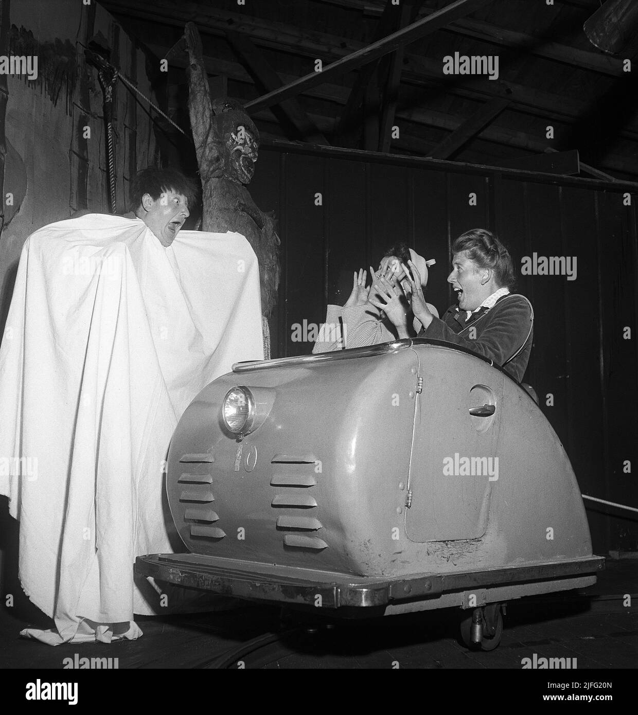 Amusement park in the 1950s. Two women are riding the scary ride and passes a man dressed as a ghost wearing a white sheet. The look surprised and afraid and are holding their hands up in horror. Sweden 1952 Kristoffersson ref Stock Photo