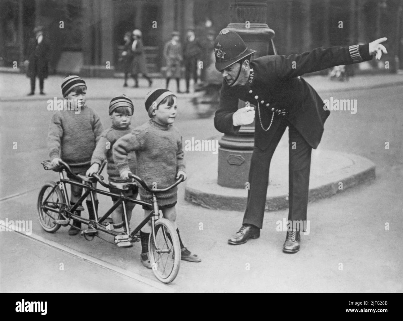 London in the 1920s. A policeman in a London street giving directions to the three children on a bicycle. The bicycle is specially made for three persons. The boys are dressed alike in woolen sweaters and caps. Stock Photo