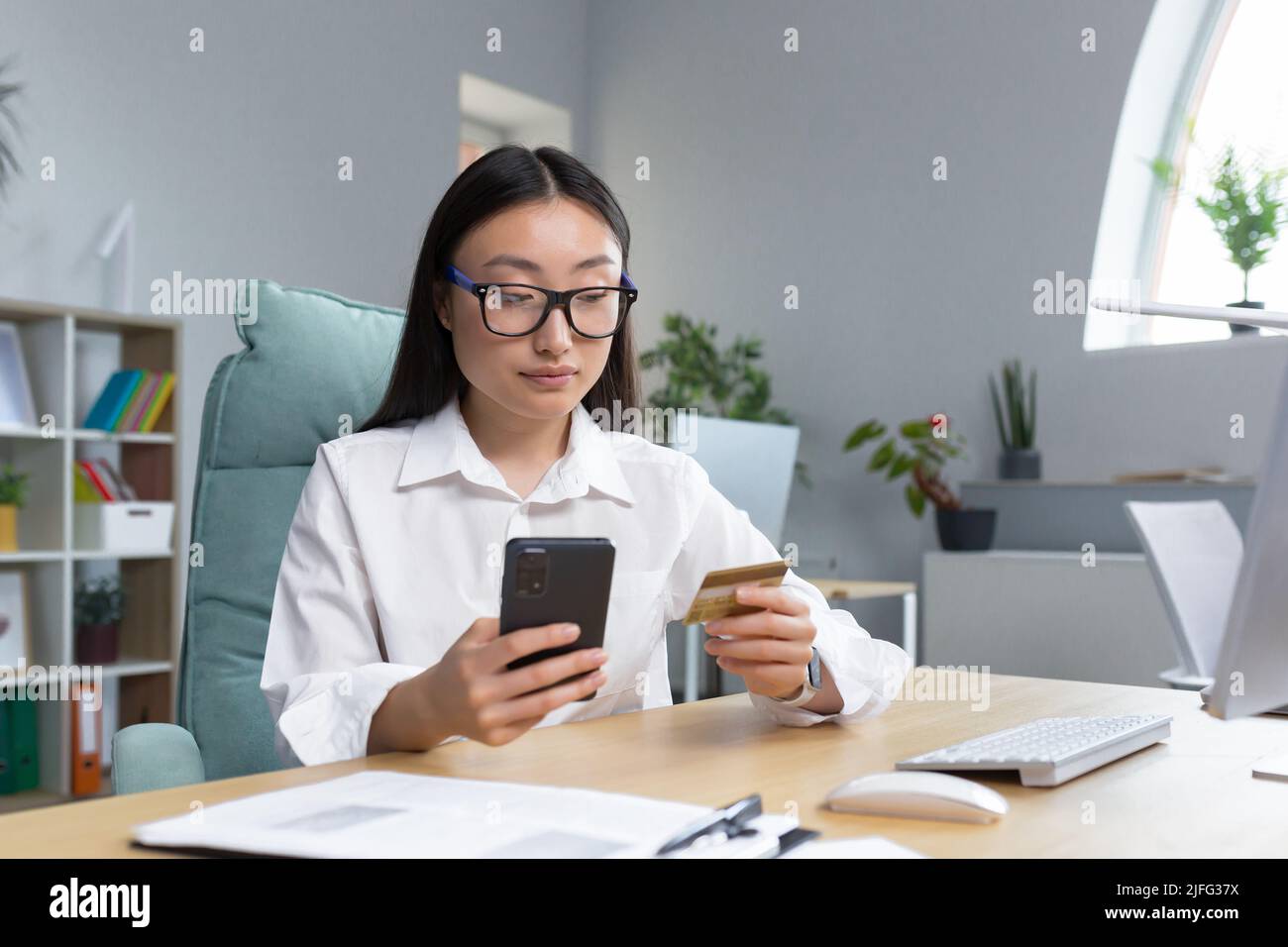Beautiful and confident Asian business woman working in the office, making banking transactions, using the phone and banking application, holding a credit card, for online shopping. Stock Photo