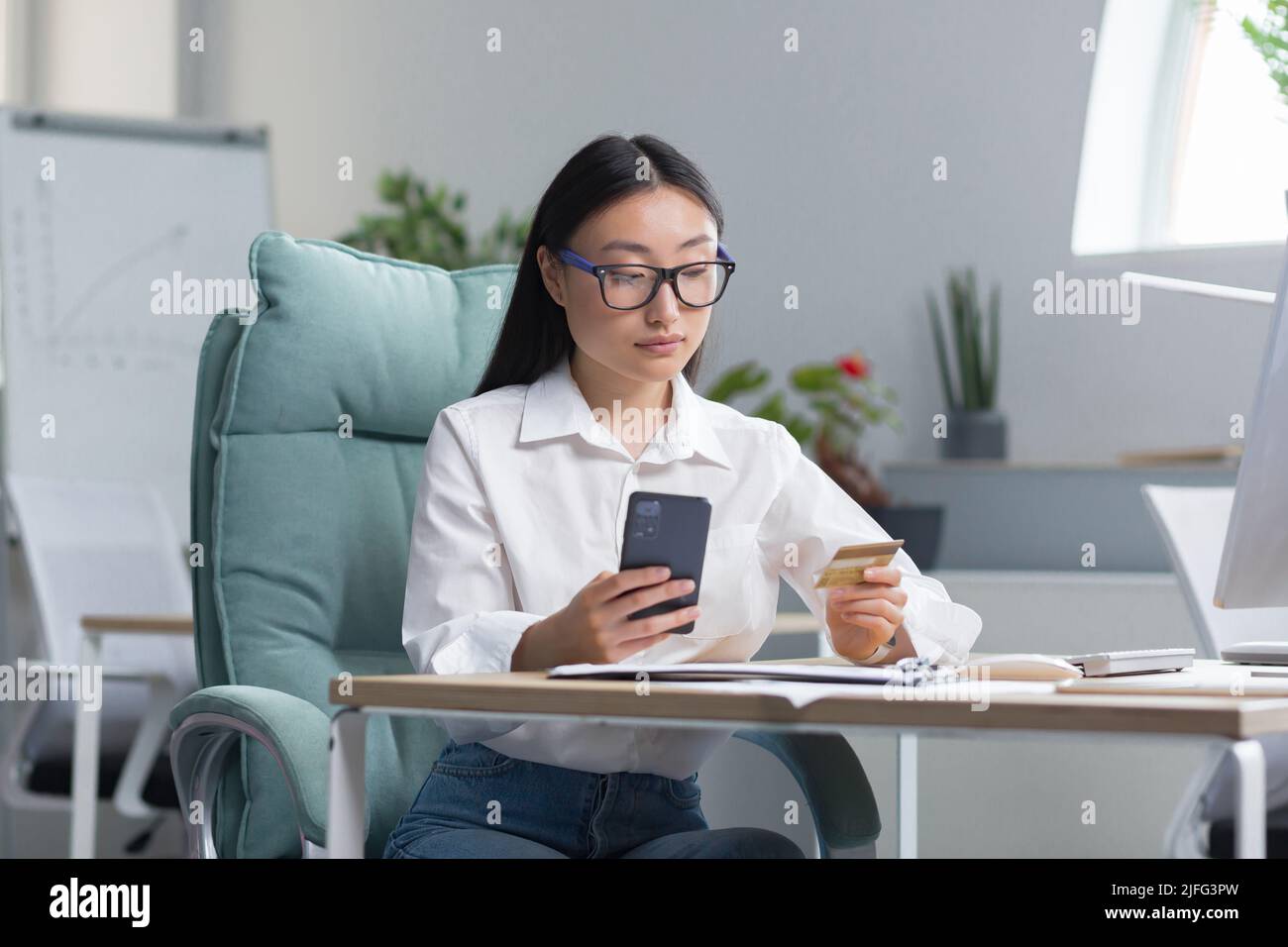 Beautiful and confident Asian business woman working in the office, making banking transactions, using the phone and banking application, holding a credit card, for online shopping. Stock Photo