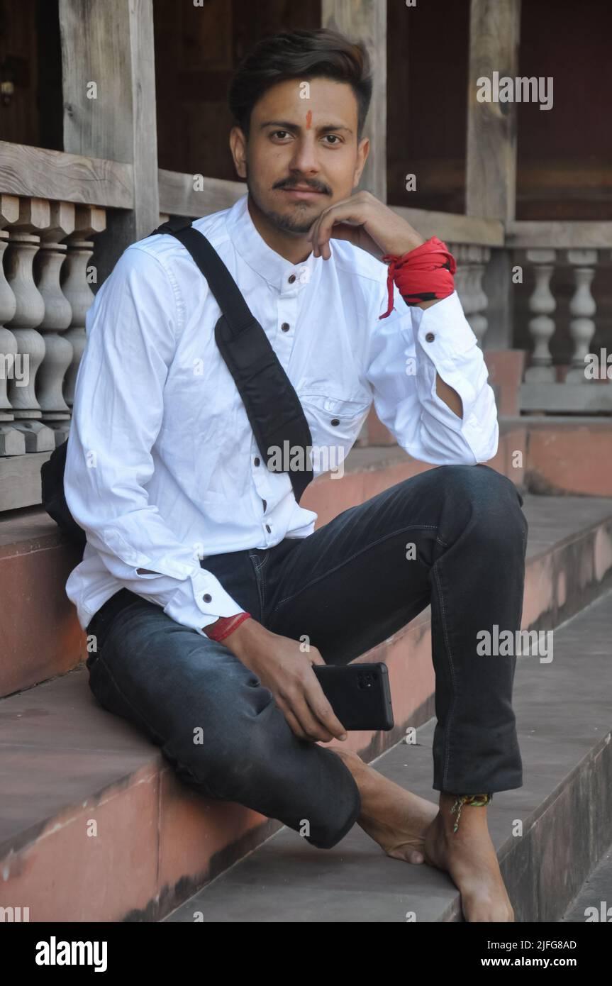 Full length of a good looking Indian young guy with looking at camera while sitting on temple stairs, posing with hand on chin and holding phone in other hand Stock Photo
