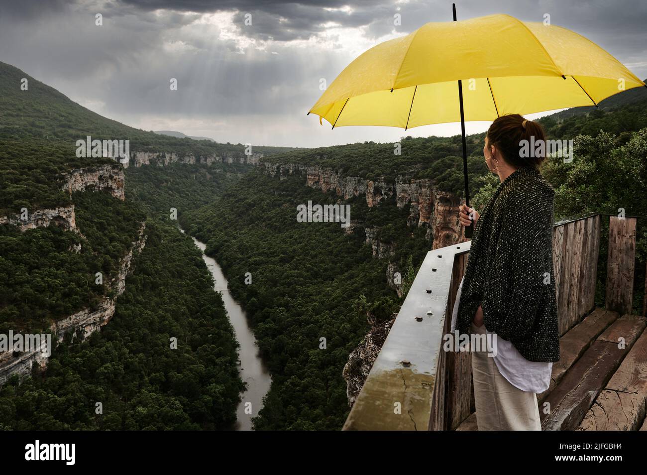Young woman observing the Ebro river bed under a yellow umbrella on a stormy afternoon, Pesquera de Ebro, Burgos, Spain Stock Photo