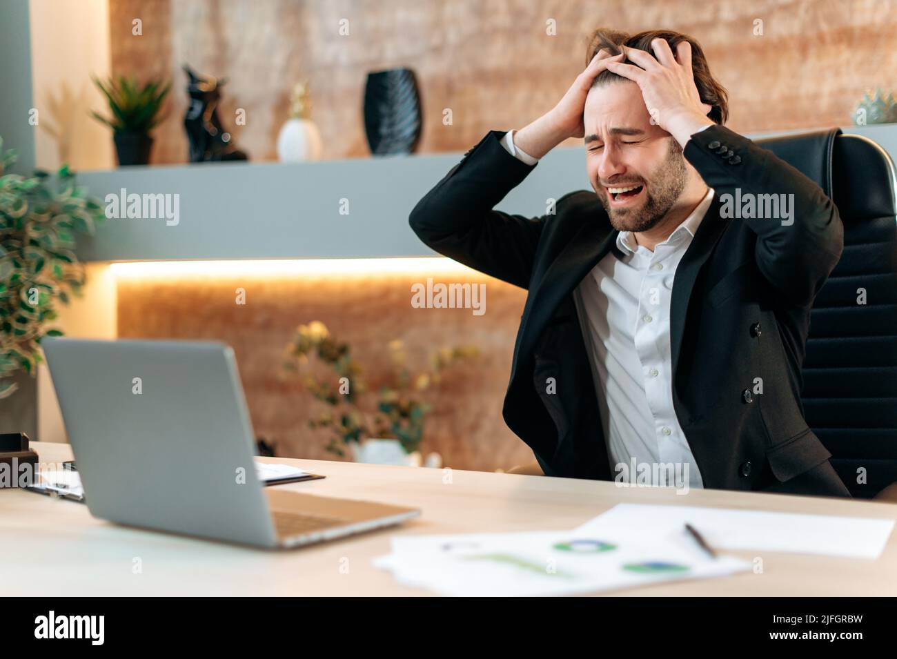 Stressed emotional caucasian businessman has problems at work, male executive, company ceo, sitting in a modern office at a desk with laptop, upset about failure, holding hands on head, annoyed Stock Photo