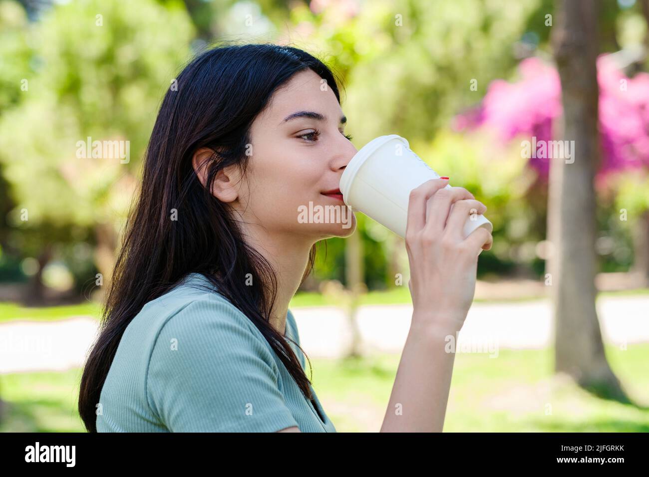 Brunette millennial woman wearing turquoise tee on city park, outdoors drinking takeaway coffee and looking into distance. Morning outdoors concept. Stock Photo