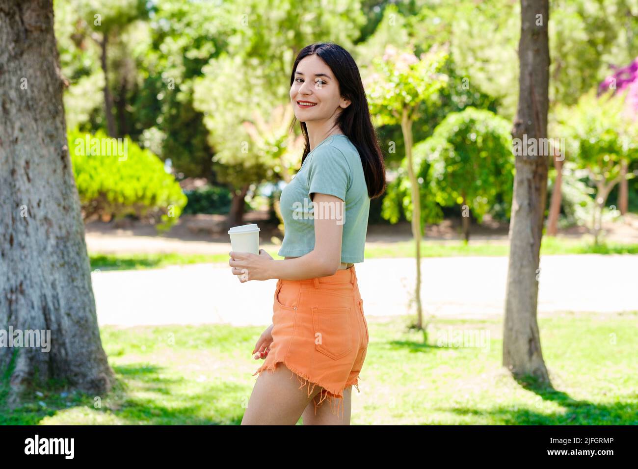 Portrait of cheerful woman wearing turquoise tee on city park, outdoors posing and looking at the camera while holding a takeaway cup of coffee. Stock Photo