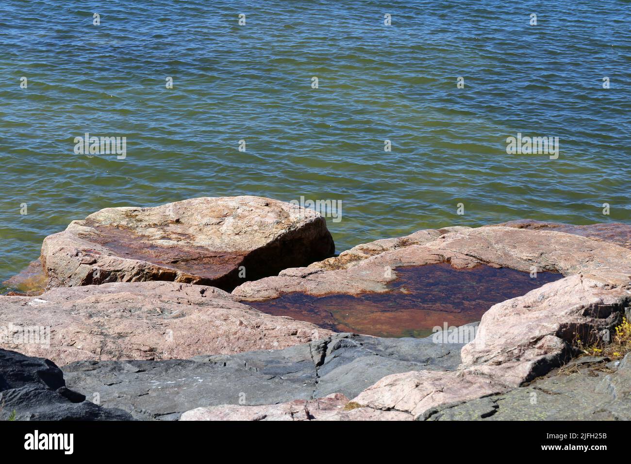 Rocks and stones meeting calm water surface of the Baltic Sea seashore in Helsinki, Finland. Sunny summer day asking you to travel, explore and swim. Stock Photo