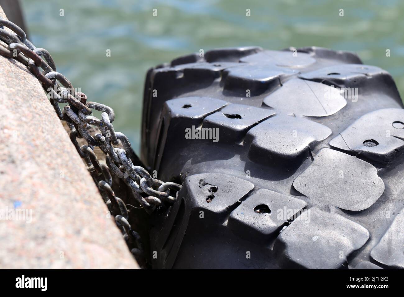 Rubber tyre used as a safety buffer for boats parking to the harbor. Maritime view with the sea, some concrete and the tire hanging with a chain. Stock Photo