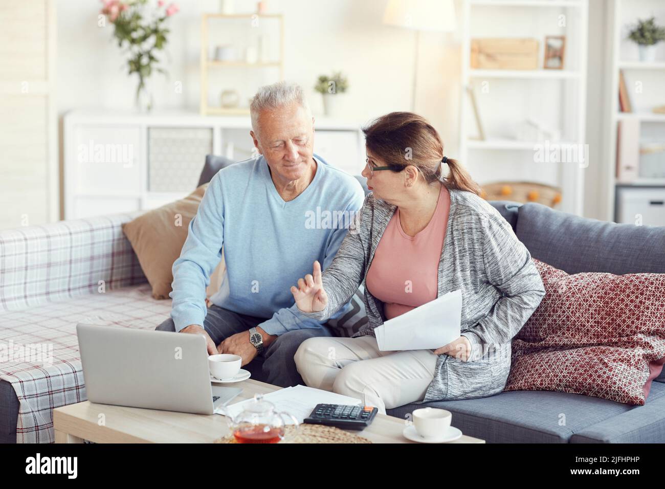 Serious busy senior couple in casual clothing sitting on sofa in living room and calculating bills while discussing family budget Stock Photo