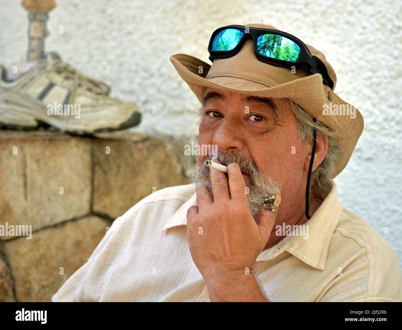 Elderly Caucasian male amputee with sad eyes sits in wheelchair and smokes cigarette, his artificial limb (leg prothesis) in the background. Stock Photo