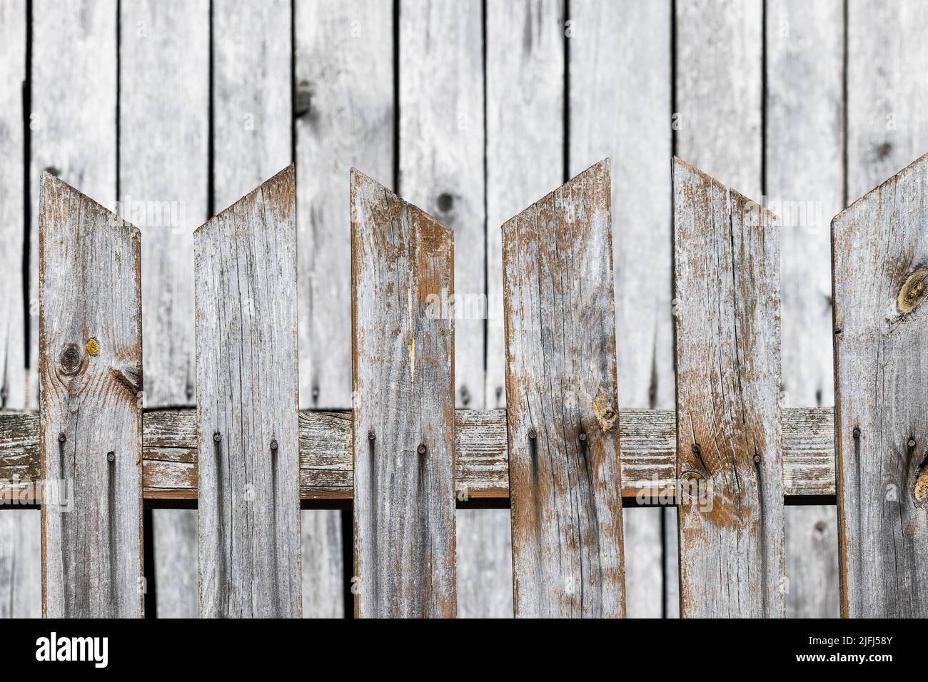 Wood fence from pointed boards with pale wooden building wall in blurred background. Closeup of weathered safety fencing with gaps between brown plank. Stock Photo