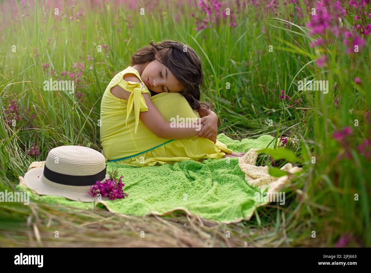 A beautiful little girl, sits hugging her knees and closing her eyes, in a yellow dress Stock Photo