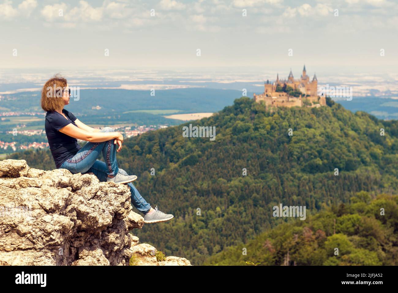 Girl tourist sitting on cliff looks at Hohenzollern Castle on mountain top, Germany. Woman travels in Swabian Alps in summer. Concept of hiking people Stock Photo