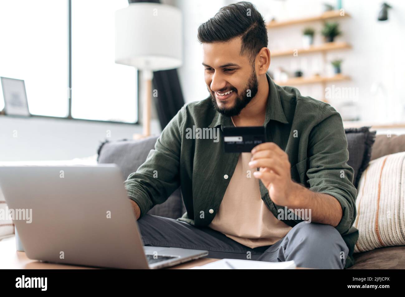 Indian or Arabian positive young male in casual wear, sitting in cozy living room, holding bank card in hand, using laptop, shopping online, ordering home delivery, paying bills, smiling Stock Photo