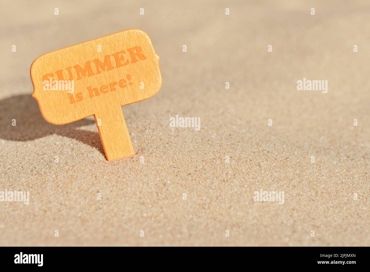 Minimal summer background, orange wooden signboard buried in the beach sand on a sunny day with text Summer is here! Warm color design with copy space Stock Photo