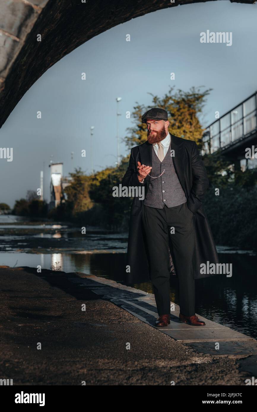 Stylish gangster man checking his pocket watch under Irish canal .1920s theme. Fashionable brutal confident bearded man. Stock Photo