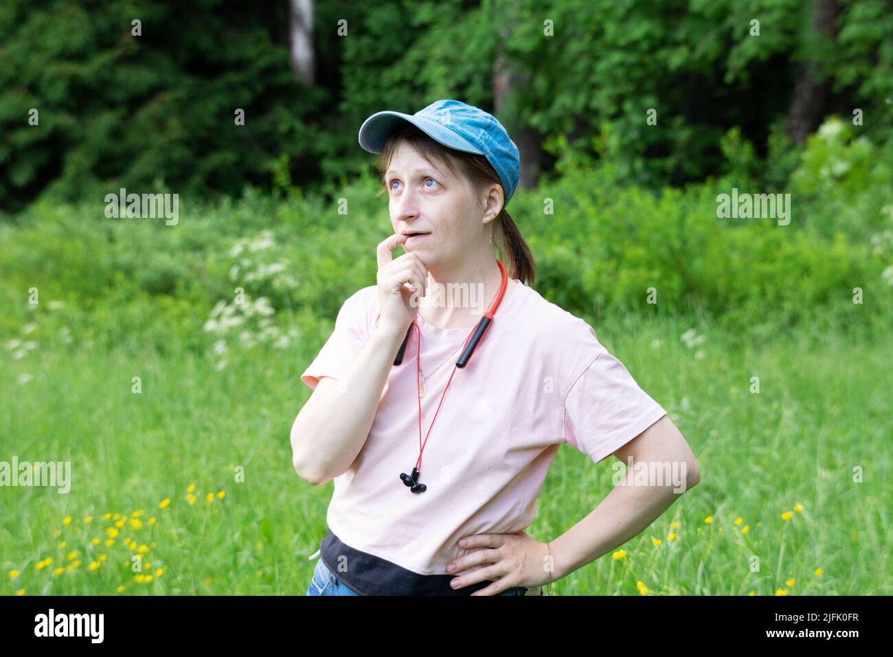 Young woman in pink shirt and jeans cap, headphones stay in forest glade and thinks. Feeling of distracted thoughtfulness Stock Photo