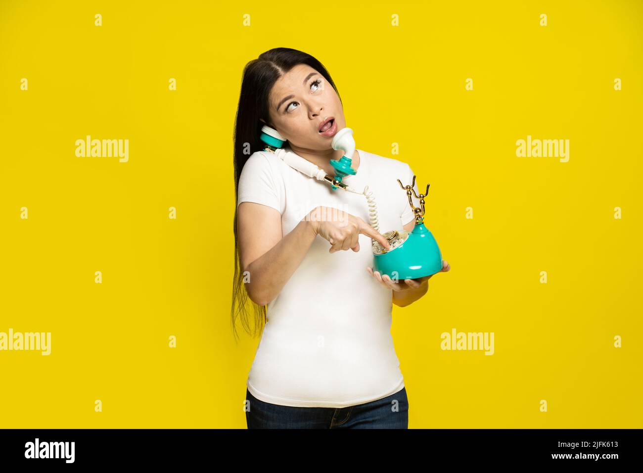 Funny asian woman using vintage, retro telephone in hands dreamingly dialling trying to remember phone number face expression wearing white t-shirt isolated on yellow background. Stock Photo