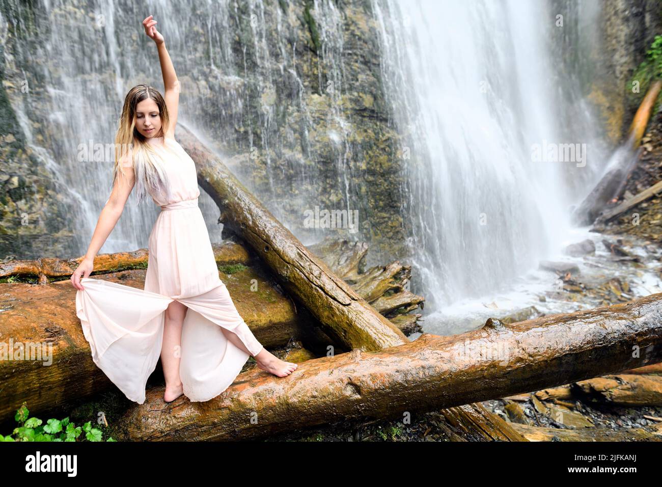 Young woman in pink dress stands barefoot on log, her arms are raised and doing some dreamlike movement and expressing happy feeling at the Bridal Stock Photo