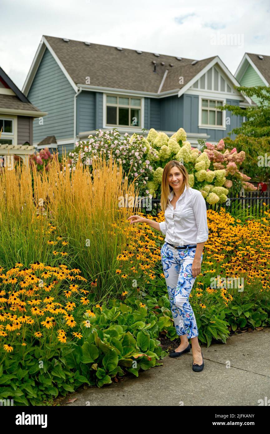 Happy attractive woman in her late 40s enjoying her beautiful flower garden and contentedly looking at her blooming hortensia and hibiscus flowers in Stock Photo