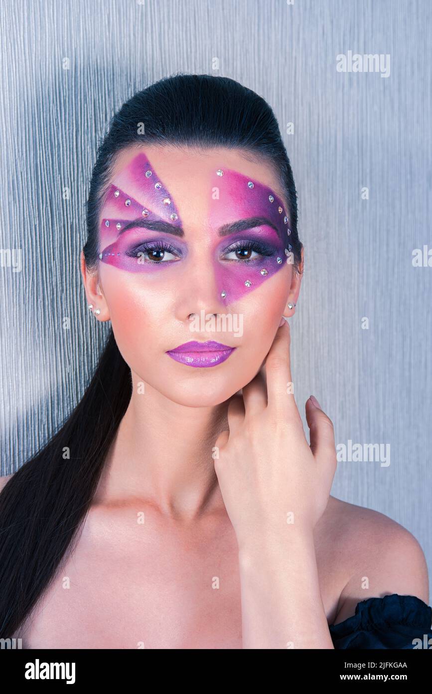 Beautiful Caucasian woman with purple face painting and crystals on her face touching her cheek and neck, on a silver background Stock Photo