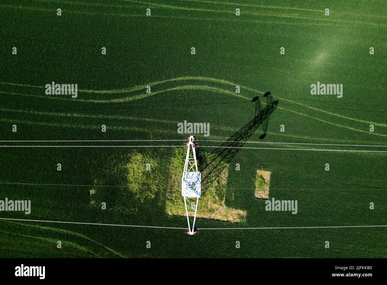 Transmission tower supporting overhead power line through cultivated crops field, aerial view from drone pov directly above Stock Photo