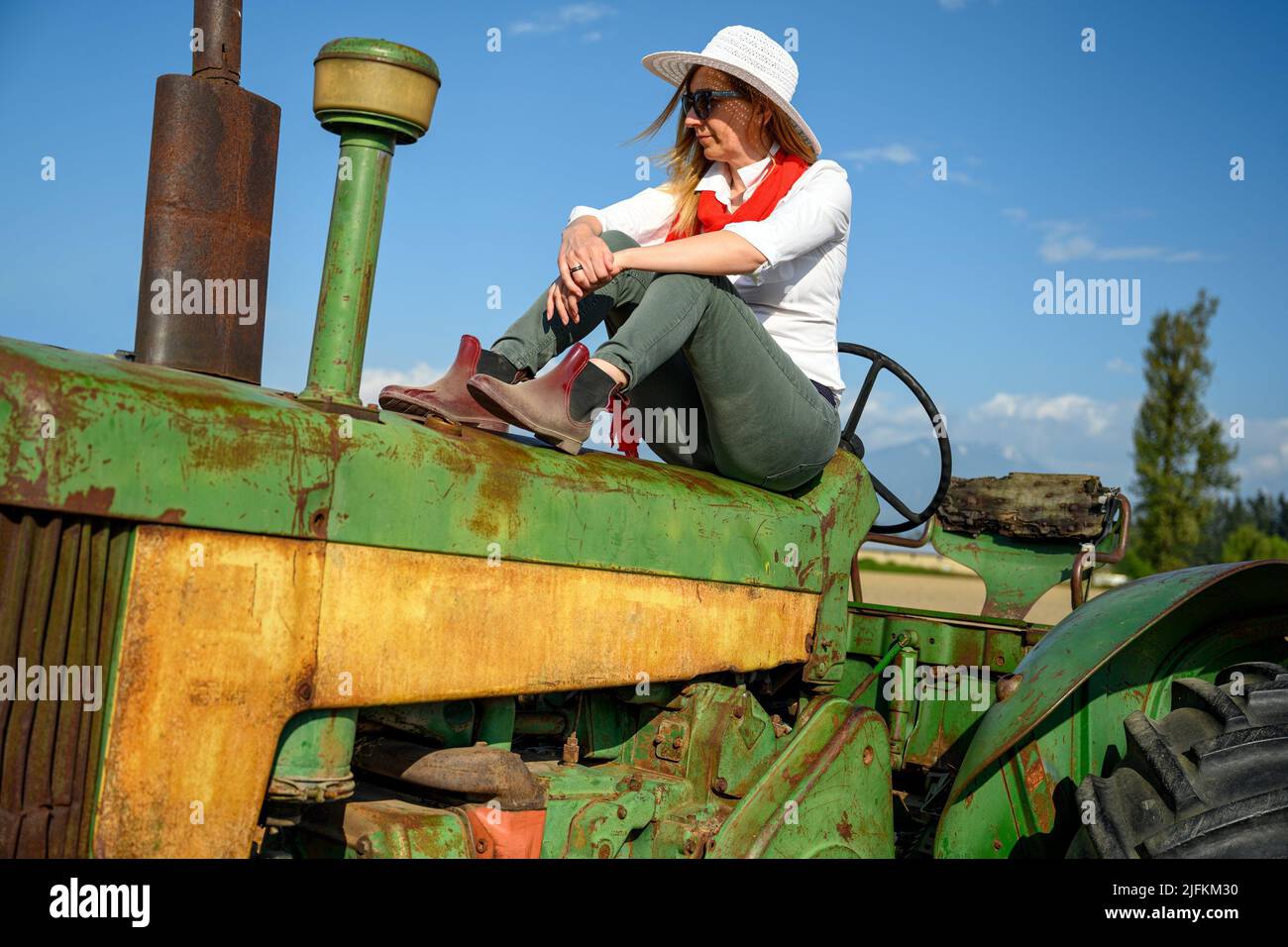 Urban woman in casual clothing sitting on an old tractor against the blue sky in the farmland. Stock Photo