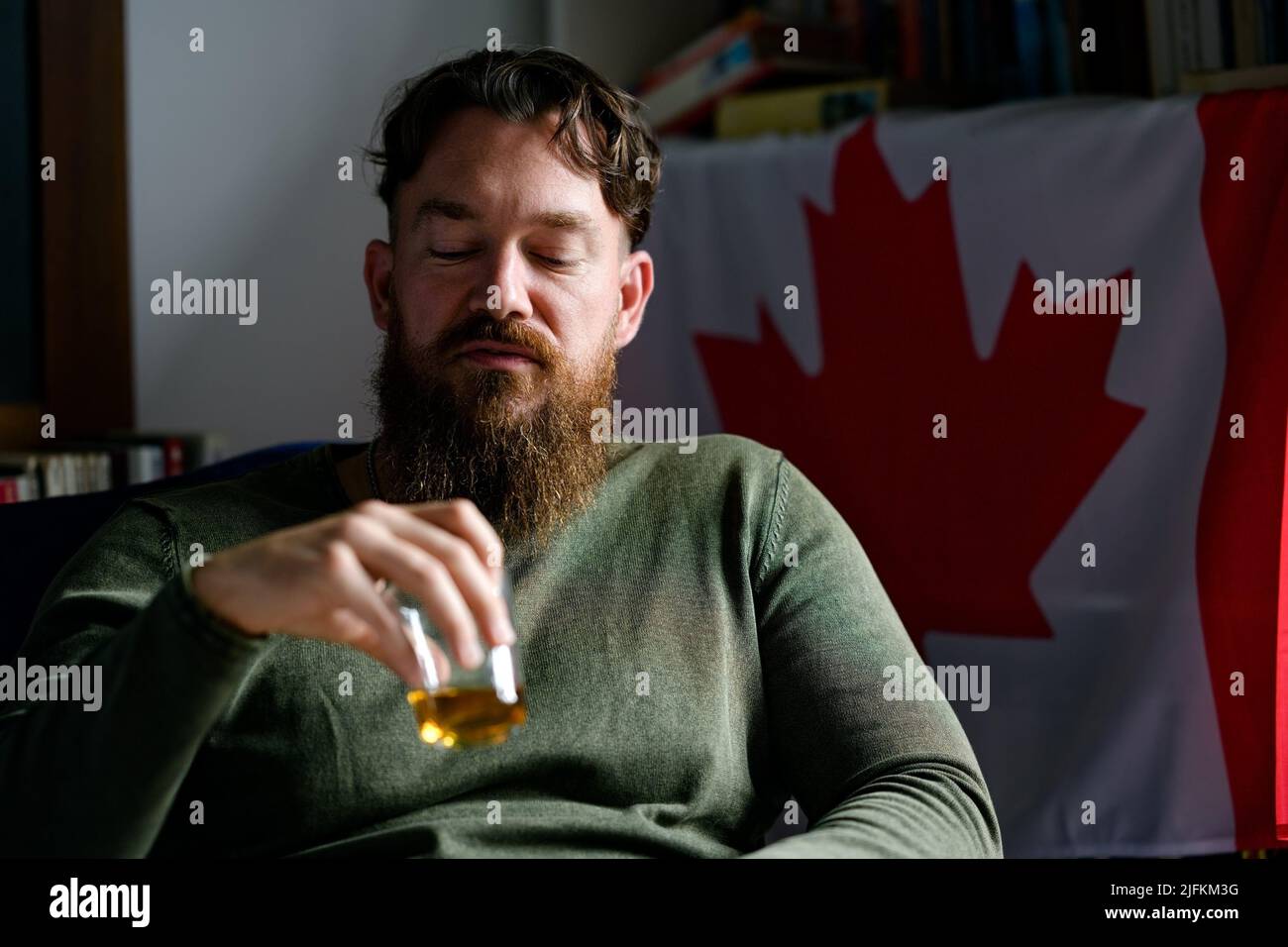 Dark tonal close up portrait of a young stylish handsome bearded Canadian man enjoying whiskey in a room with a Canadian flag behind him. Stock Photo