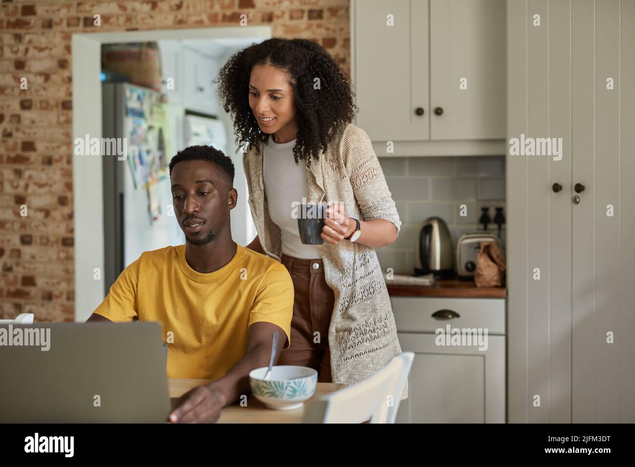 Young multiethnic couple talking and working on a laptop while drinking coffee together in their kitchen in the morning Stock Photo