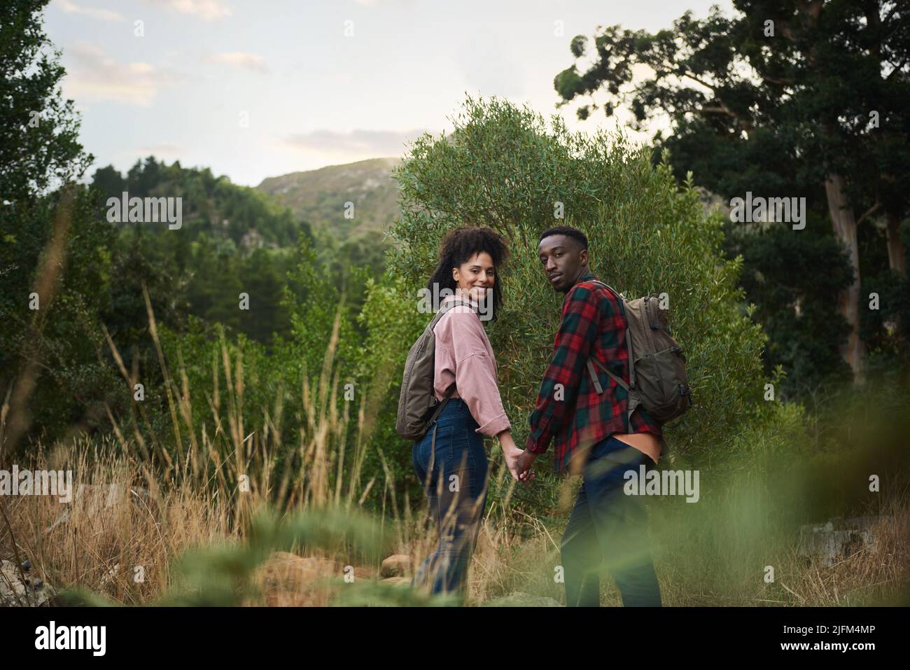 Portrait of a smiling young multiethnic couple hiking hand in hand together along a path in some rugged hills in the summertime Stock Photo