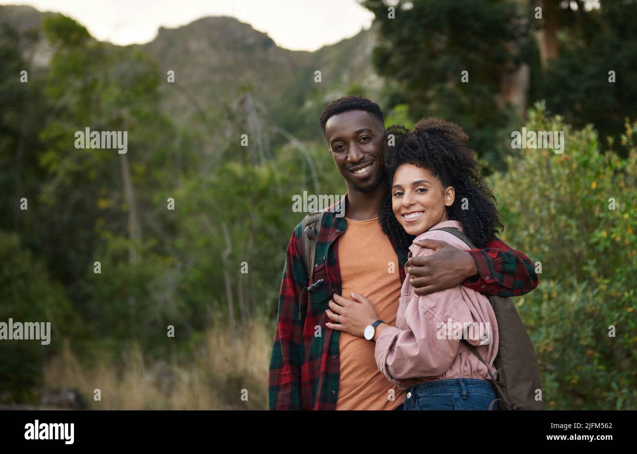 Portrait of smiling young multiethnic couple standing arm in arm on a trail during a hike together in some rugged hills in the summertime Stock Photo
