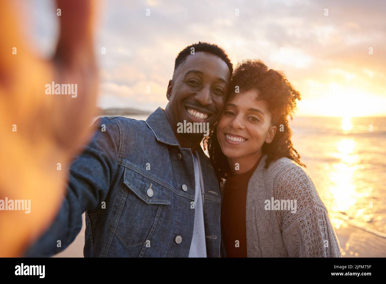 Loving young multiethnic couple smiling and taking a selfie while standing cheek to cheek on a beach at sunset Stock Photo