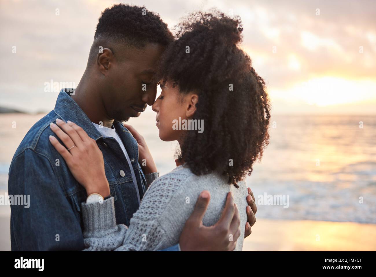 Affectionate young multiethnic couple standing face to face with their eyes closed in each other's arms on a beach at sunset Stock Photo