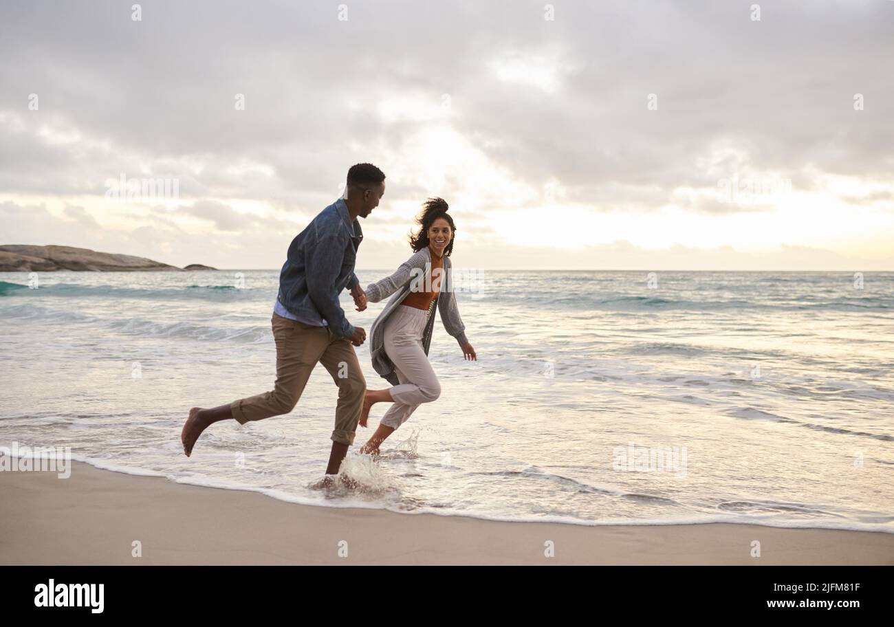Carefree young multiethnic couple laughing while running hand in hand together through the water on a beach on an overcast afternoon Stock Photo