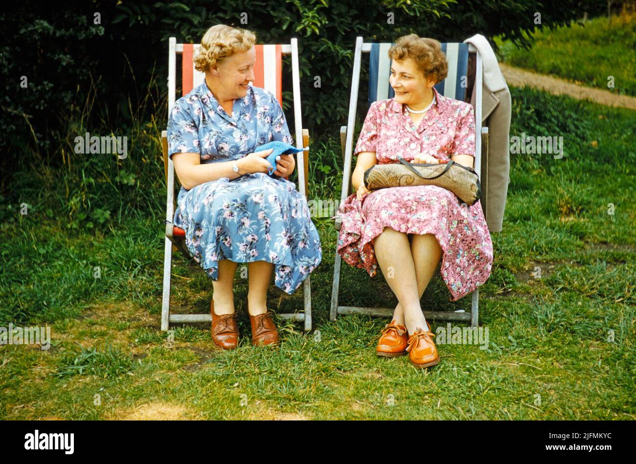 Two women friends sitting in deckchairs on grass lawn one knitting, smiling at each other,  England, UK early 1960s Stock Photo