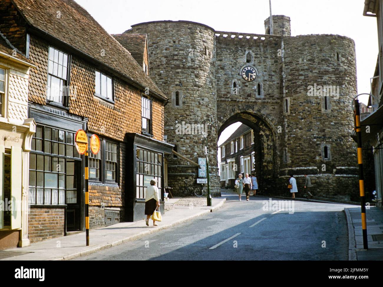 People walking in street by historic Landgate Gateway arched gatehouse,, Rye, Kent, England, UK early 1960s Stock Photo