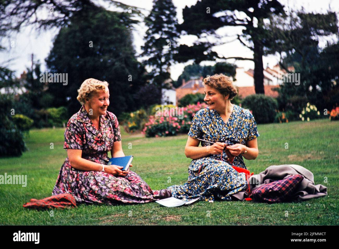 Two women friends sitting on grass lawn one knitting, smiling at each other, Dawlish, Devon, England, UK early 1960s Stock Photo
