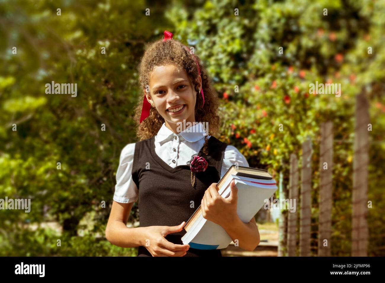 Portrait of a beautiful smiling mixed raced Latin young girl holding her school books in her arms, she is standing outdoors under the direct sun light Stock Photo