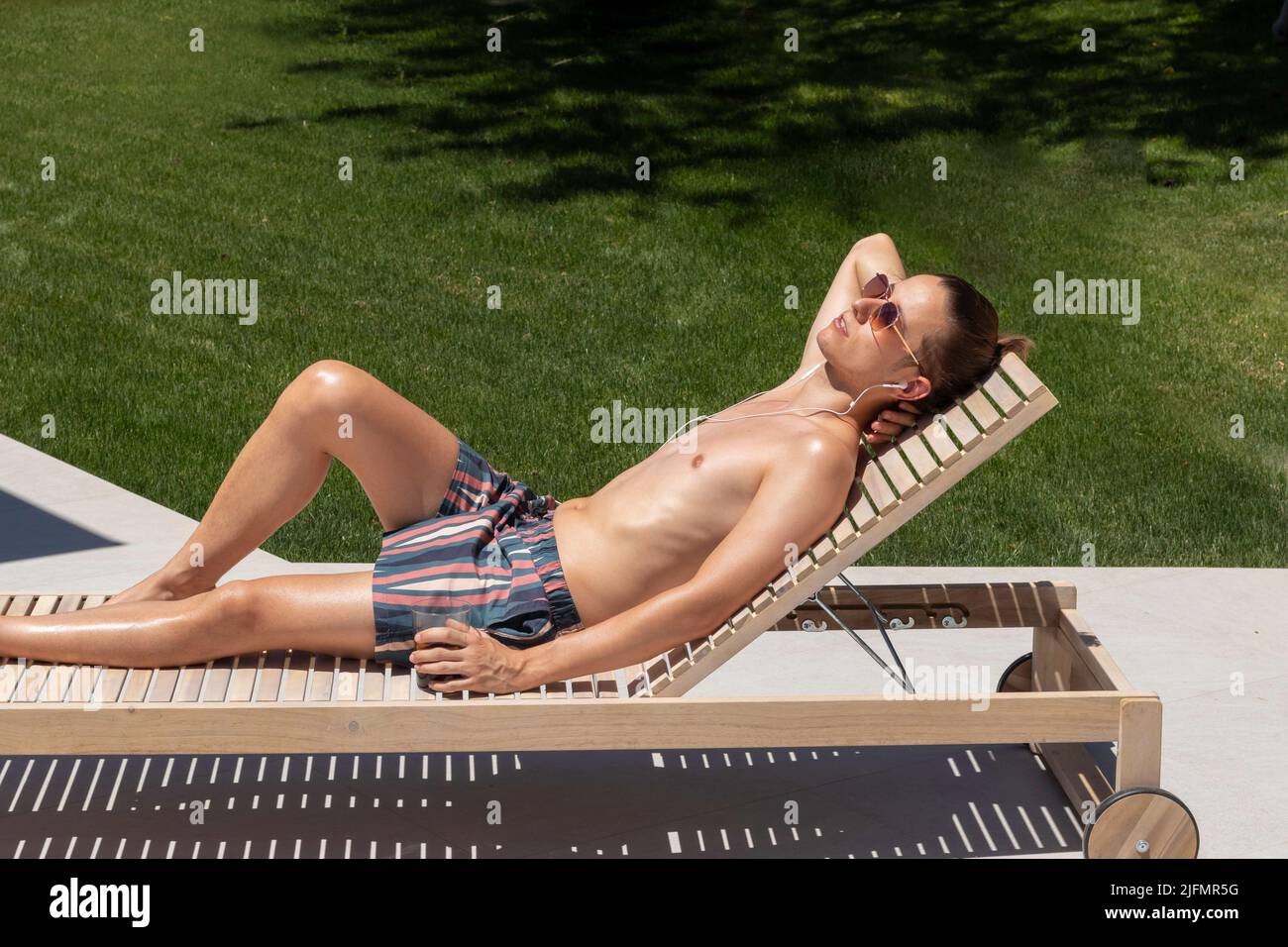 Young man on sunglasses and swimsuit lying on a sun lounger sun bathing close to the garden grass in summer time Stock Photo