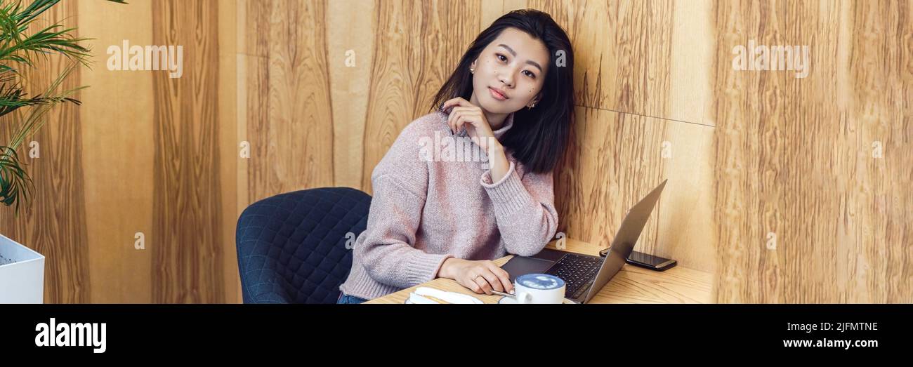 Cute adult girl plays in a laptop in a coffee shop. Asian woman working in a cafe at a laptop. Freelance concept. Web banner Stock Photo