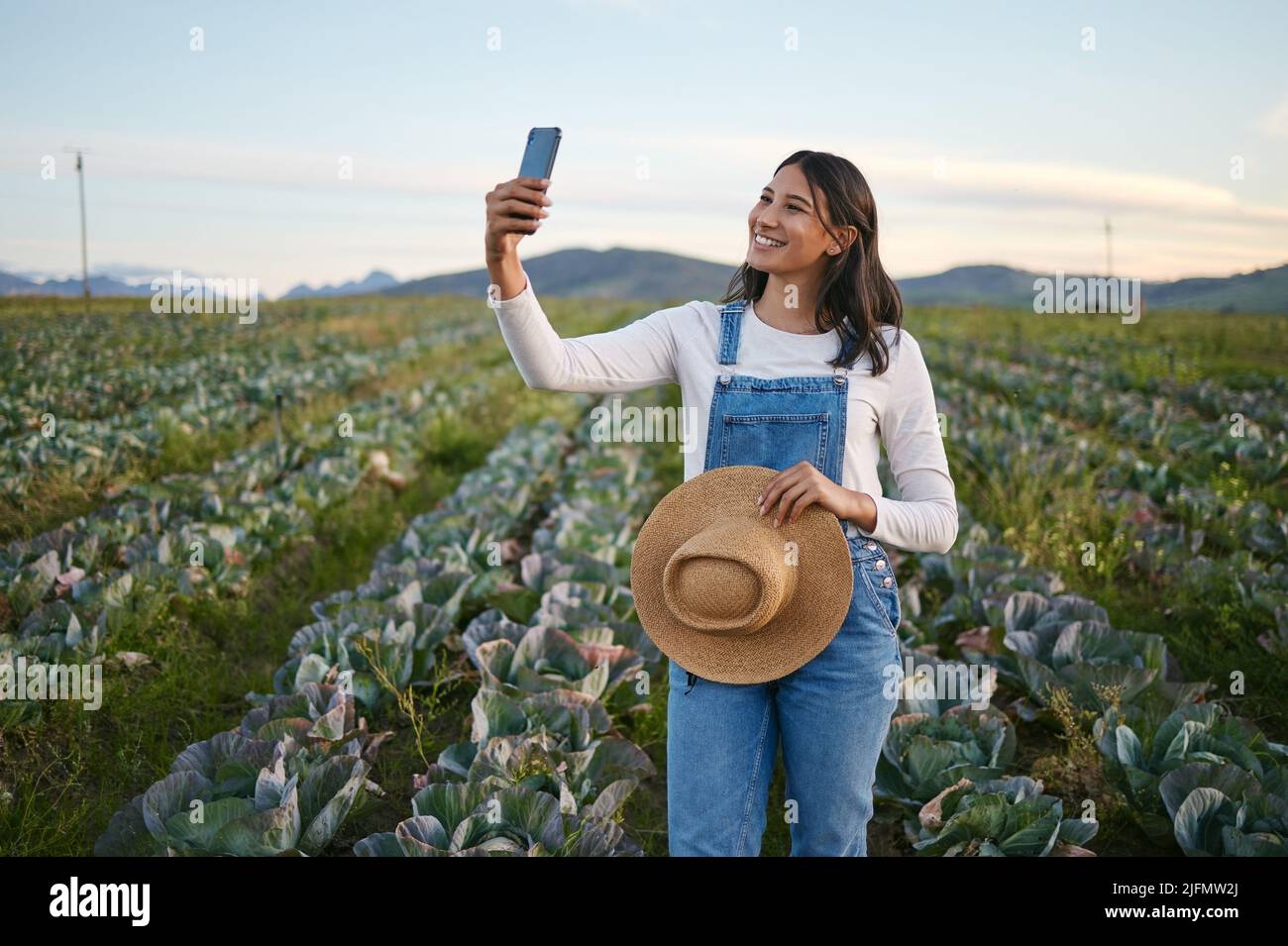 Woman farmer taking a selfie on her smartphone while standing in a cabbage field. Young brunette female with a straw hat using her mobile device on an Stock Photo
