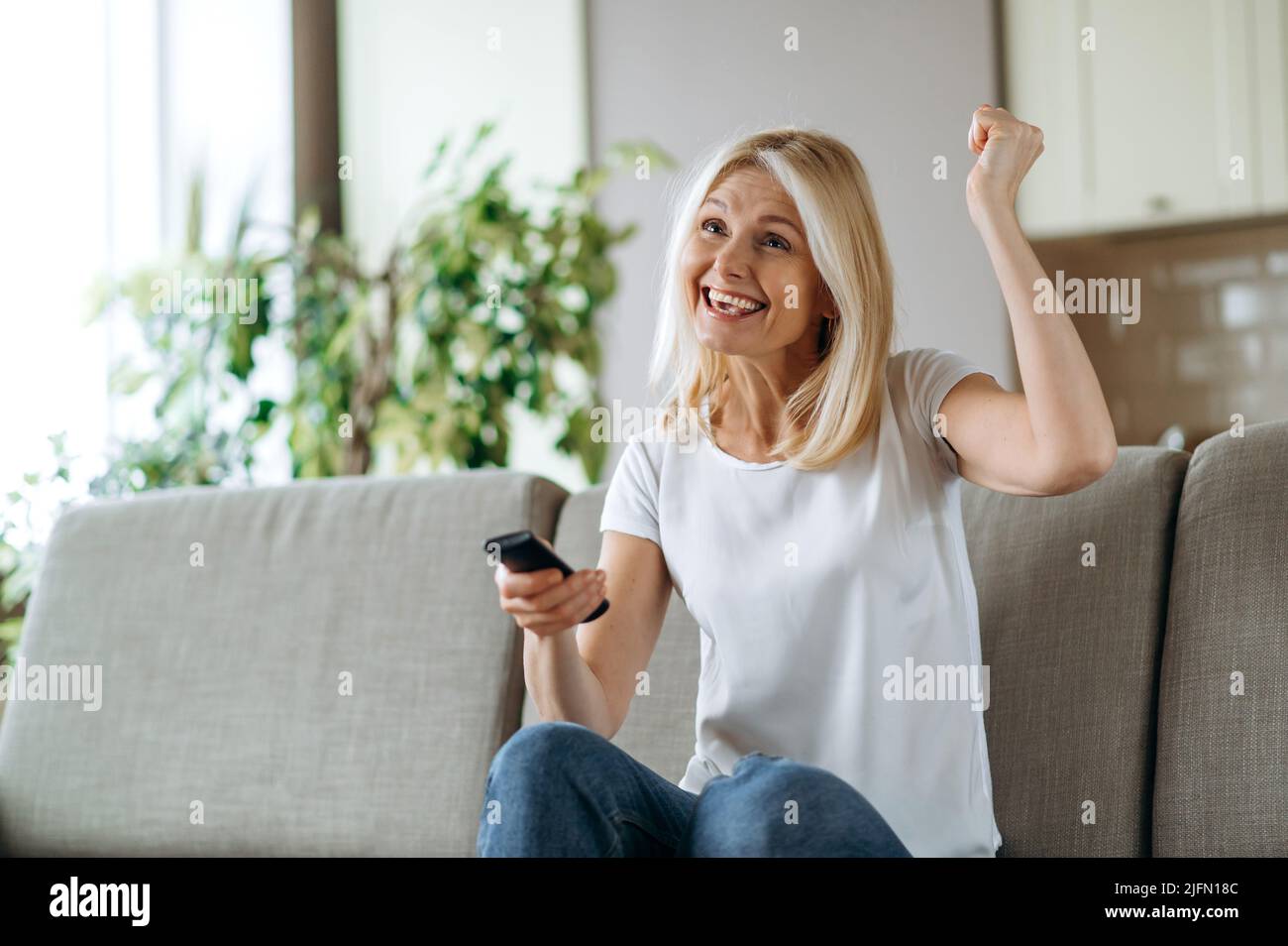 Leisure and entertainment concept. Positive excited caucasian blonde woman watching TV show or film, holding remote control, changes channels, while sitting on cozy sofa at home in living room, smiles Stock Photo